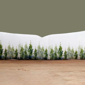 May include: A white lumbar pillow featuring a watercolor design of a forest of evergreen trees in shades of green and blue. The pillow is rectangular and rests on a wooden surface against a muted green background.
