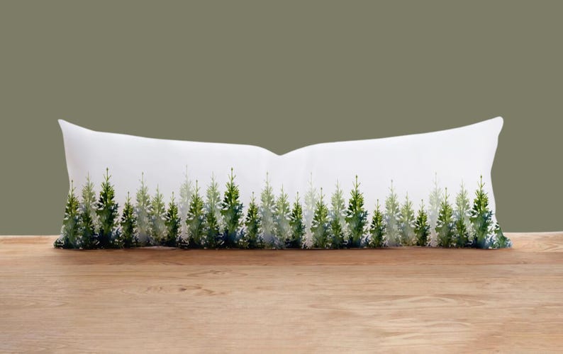 May include: A white lumbar pillow featuring a watercolor design of a forest of evergreen trees in shades of green and blue. The pillow is rectangular and rests on a wooden surface against a muted green background.