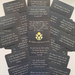 May include: A set of gray oracle cards with inspirational messages. The cards feature a variety of phrases such as "I will enjoy the connections with people who love, cherish and value me." and "I am ready to level up. My next steps will be?" The cards are arranged in a random pattern on a dark surface.
