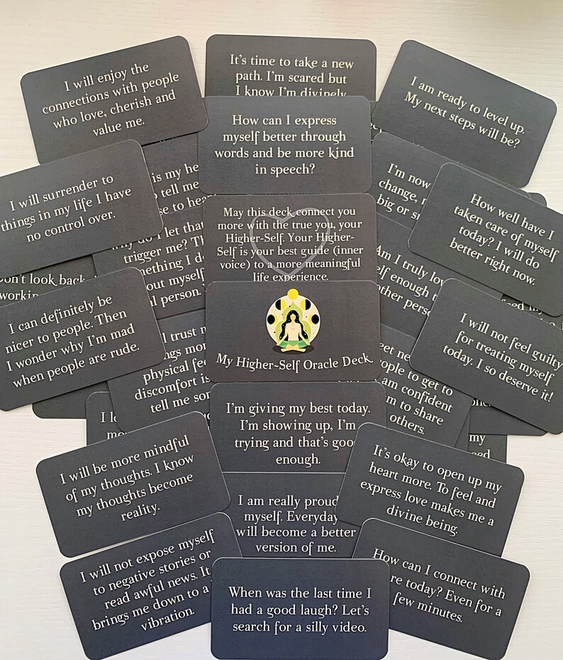 May include: A set of gray oracle cards with inspirational messages. The cards feature a variety of phrases such as "I will enjoy the connections with people who love, cherish and value me." and "I am ready to level up. My next steps will be?" The cards are arranged in a random pattern on a dark surface.