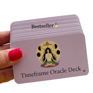 May include: A deck of Timeframe Oracle cards held by a hand with a bright pink nail. The cards are light purple with the word "Bestseller" and a gold medal icon. The deck features a circular logo with a meditating figure and the words "Total Divine".