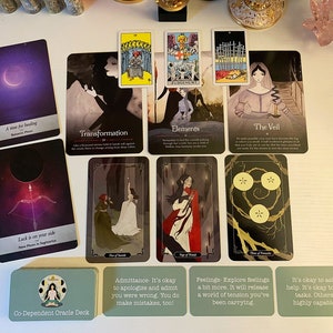 May include: A spread of tarot and oracle cards on a white surface. The cards feature illustrations of people, symbols, and landscapes. The cards are arranged in a grid pattern. The text on the cards includes phrases like "Transformation", "Elements", "The Veil", "Luck is on your side", "Co-Dependent Oracle Deck", "Admittance- It's okay to apologize and admit you were wrong. You do make mistakes, too!", "Feelings-Explore feelings a bit more. It will release a world of tension you've been carrying", and "It's okay to ask for help. It's okay to share tasks. Others are highly capable too."