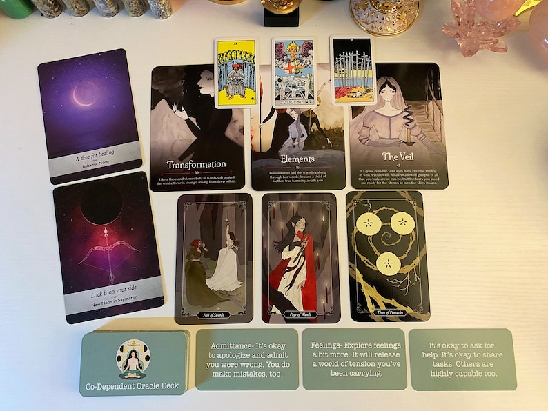 May include: A spread of tarot and oracle cards on a white surface. The cards feature illustrations of people, symbols, and landscapes. The cards are arranged in a grid pattern. The text on the cards includes phrases like "Transformation", "Elements", "The Veil", "Luck is on your side", "Co-Dependent Oracle Deck", "Admittance- It's okay to apologize and admit you were wrong. You do make mistakes, too!", "Feelings-Explore feelings a bit more. It will release a world of tension you've been carrying", and "It's okay to ask for help. It's okay to share tasks. Others are highly capable too."