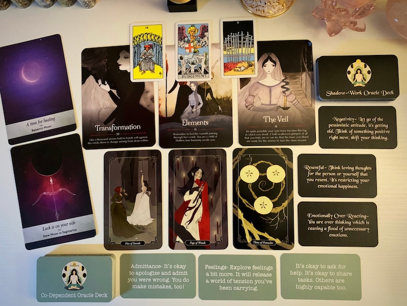 May include: A spread of tarot and oracle cards on a white surface. The cards feature illustrations of people, symbols, and landscapes. The cards are arranged in a grid pattern. The cards are from the Shadow-Work Oracle Deck, Co-Dependent Oracle Deck, and a standard tarot deck.
