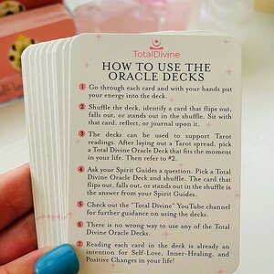 May include: A close-up of a set of oracle cards with instructions on how to use them. The cards are cream-colored with pink and gold accents. The text on the cards reads "Total Divine" and "How to Use the Oracle Decks".