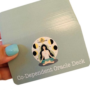May include: A deck of cards with a light blue background and a white border. The front of the card features a woman meditating with a moon and stars design. The text "Co-Dependent Oracle Deck" is printed in white on the bottom of the card.