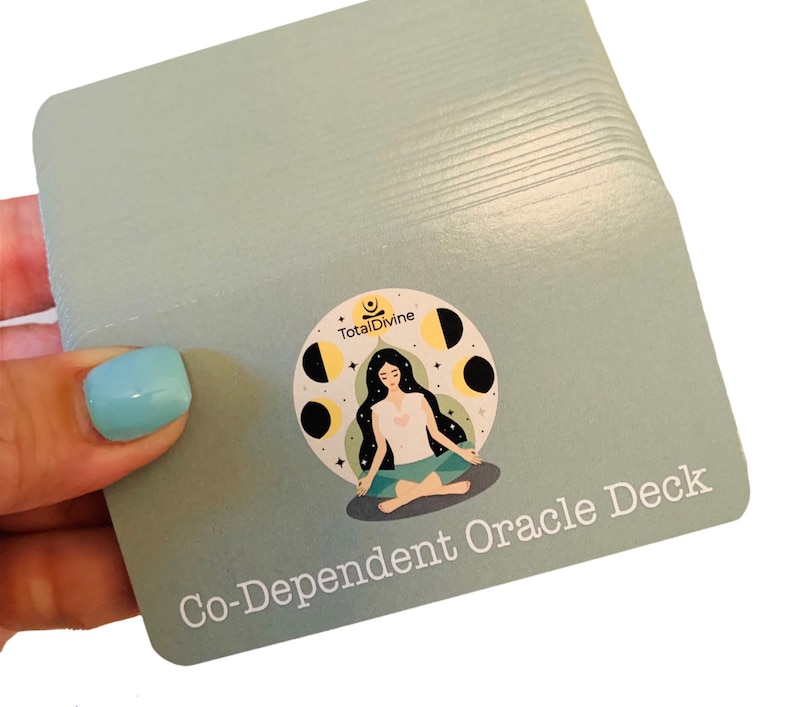 May include: A deck of cards with a light blue background and a white border. The front of the card features a woman meditating with a moon and stars design. The text "Co-Dependent Oracle Deck" is printed in white on the bottom of the card.
