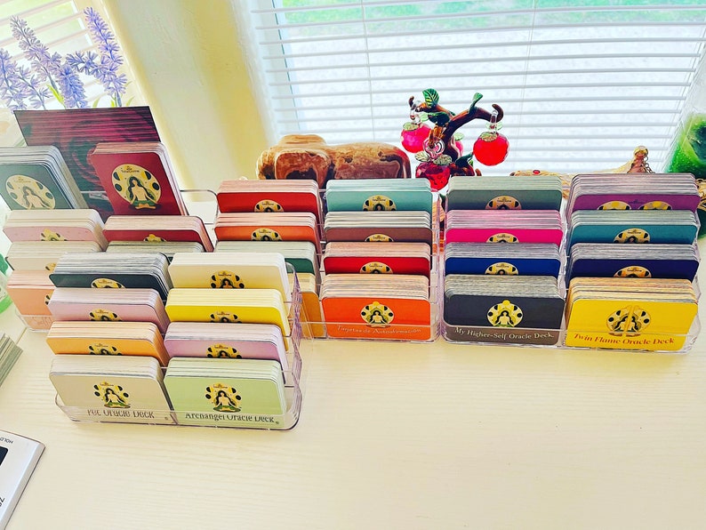 May include: A collection of colorful oracle decks organized in clear plastic holders. The decks are labeled with their names, including "My Higher Self Oracle Deck", "Twin Flame Oracle Deck", "Archangel Oracle Deck", and "Full Moon Oracle Deck".