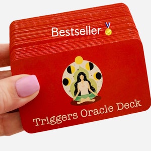 May include: A red deck of cards with the title "Triggers Oracle Deck" and a woman meditating in a circle of moons. The deck is labeled "Bestseller" with a gold medal.