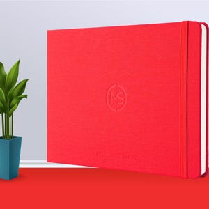 May include: A red hardback notebook with a white elastic band closure and a small embossed logo on the front. The notebook is on a red surface.