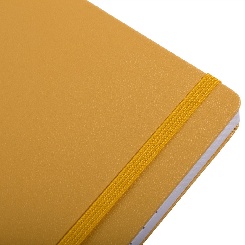 May include: Close-up of a mustard-colored notebook with a matching elastic band closure. The cover has a textured, leather-like appearance. The pages are white with a subtle dotted line.