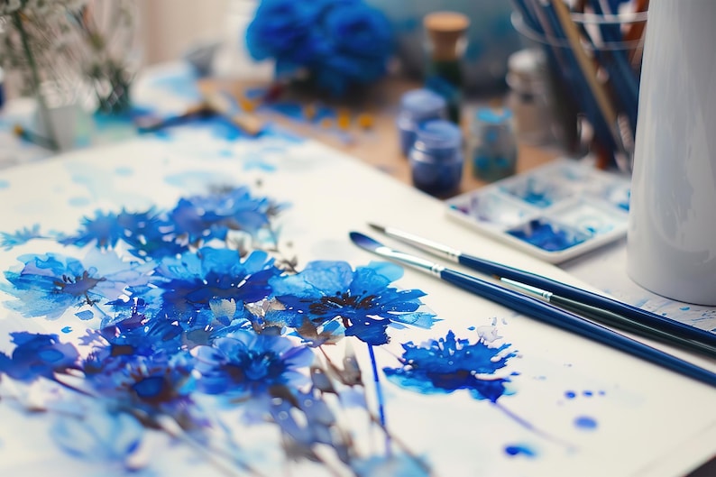 May include: A watercolour painting of blue flowers on white paper. Two blue paintbrushes are lying on the paper.