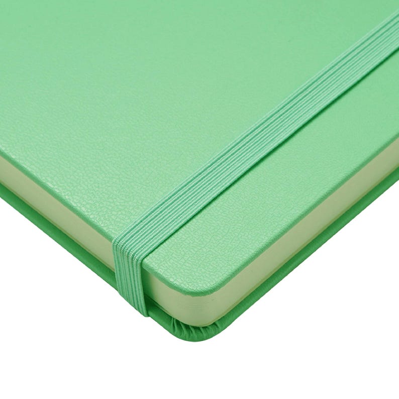 Puede incluir: Primer plano de un cuaderno verde menta con un cierre de banda el&aacute;stica a juego. El cuaderno tiene una cubierta texturizada y p&aacute;ginas de color crema. La banda el&aacute;stica se extiende por la cubierta del cuaderno.