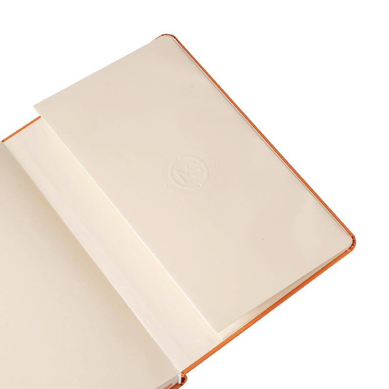 Puede incluir: Un cuaderno abierto, encuadernado en naranja, con p&aacute;ginas color crema. La p&aacute;gina superior derecha tiene un dise&ntilde;o sutil en relieve. Las p&aacute;ginas del cuaderno son ligeramente blanquecinas y la encuadernaci&oacute;n es de color naranja c&aacute;lido.