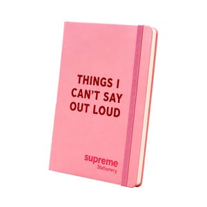 May include: A pink hardback notebook with an elastic closure band. The front cover features the dark pink text "THINGS I CAN'T SAY OUT LOUD" and the "supreme Stationery" logo printed in the bottom left corner. The notebook has white pages.