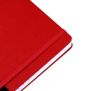May include: A red notebook with a textured cover and a matching elastic band closure. The notebook's pages are white with a dotted pattern. The design is simple and classic.