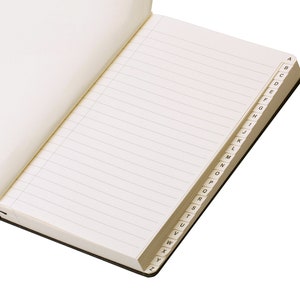 Pode incluir: Um caderno aberto, com linhas, com capa preta e um &iacute;ndice alfab&eacute;tico na borda das p&aacute;ginas.