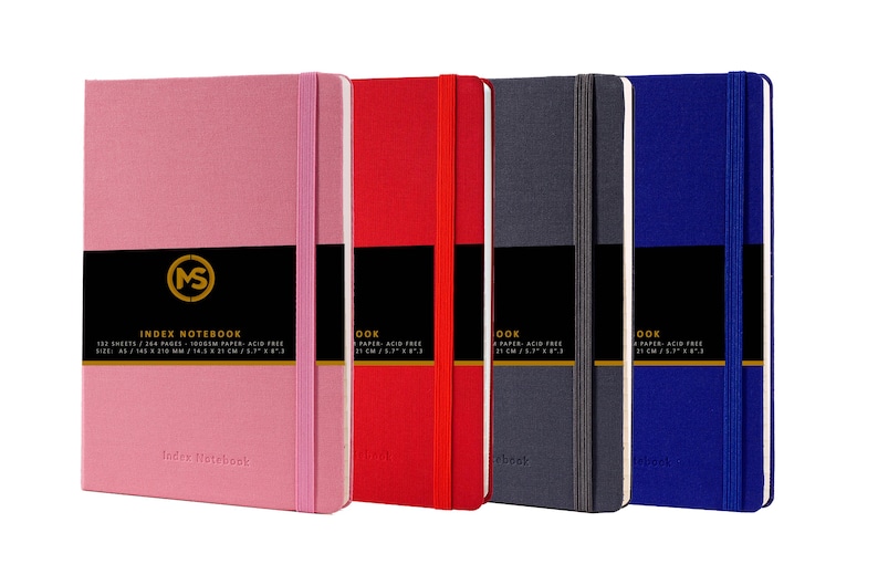 Pode incluir: Quatro cadernos de &iacute;ndice de capa dura em rosa, vermelho, cinza e azul. Cada caderno tem uma faixa preta com letras douradas que dizem "INDEX NOTEBOOK" e um logotipo dourado. Os cadernos est&atilde;o todos fechados e alinhados em uma fila.