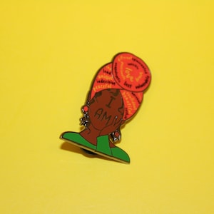 May include: A red and orange enamel pin featuring a black woman with a headscarf. The headscarf is covered in words, including "love", "unique", "brave", and "powerful". The woman is holding her hand to her cheek and the words "I AM" are written across her face.
