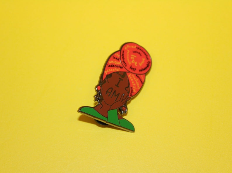 May include: A red and orange enamel pin featuring a black woman with a headscarf. The headscarf is covered in words, including "love", "unique", "brave", and "powerful". The woman is holding her hand to her cheek and the words "I AM" are written across her face.
