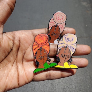 May include: Three enamel pins featuring a cartoon illustration of a woman with a headscarf. Each pin has a circle around the woman's head with the words "I AM" in the center and positive affirmations around the circle. The first pin has a red, orange, and yellow circle with the words "love, brave, unique, peaceful, confident, beautiful, lost, passionate, invisible, opinionated, vulnerable". The second pin has a pink, orange, and yellow circle with the words "love, brave, unique, peaceful, confident, beautiful, lost, passionate, invisible, opinionated, vulnerable". The third pin has a light blue, pink, and yellow circle with the words "love, brave, unique, peaceful, confident, beautiful, lost, passionate, invisible, opinionated, vulnerable".