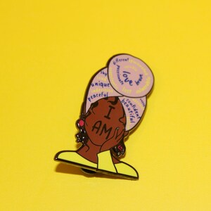 May include: A pink and yellow enamel pin featuring a cartoon illustration of a woman with a headscarf. The headscarf has words written on it, including "love", "unique", "confident", and "beautiful". The woman is wearing a yellow shirt and has a red earring. The pin says "I AM" in large letters.