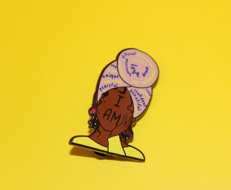 May include: A pink and yellow enamel pin featuring a cartoon illustration of a woman with a headscarf. The headscarf has words written on it, including "love", "unique", "confident", and "beautiful". The woman is wearing a yellow shirt and has a red earring. The pin says "I AM" in large letters.