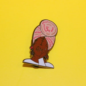 May include: A gold-toned enamel pin featuring a cartoon illustration of a Black woman with a pink and white head wrap. The head wrap has words written on it, including "I am", "love", "unique", "confident", and "beautiful".