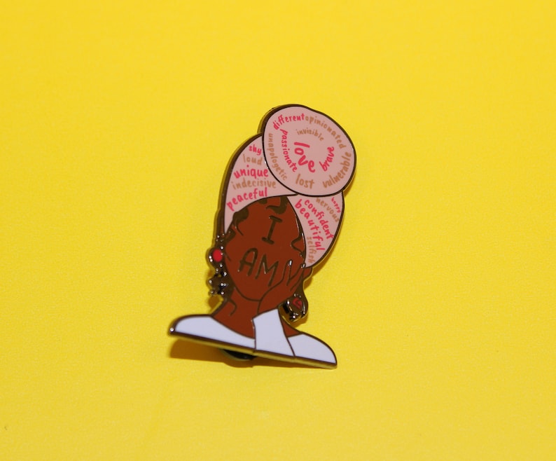May include: A gold-toned enamel pin featuring a cartoon illustration of a Black woman with a pink and white head wrap. The head wrap has words written on it, including "I am", "love", "unique", "confident", and "beautiful".