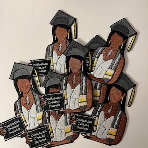 May include: A set of six black and gray graduation patches featuring a cartoon image of a Black woman wearing a graduation cap and gown. Each patch has a white and yellow striped sash and a black sign that reads "Dedication Created Success".