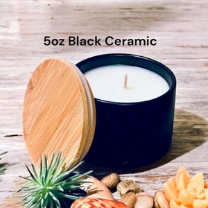 May include: A 5 oz black ceramic candle with a white wax top and a wooden lid. The candle is sitting on a wooden surface with small rocks and a succulent plant.