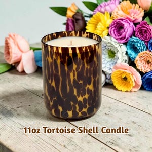 May include: A cylindrical candle in a tortoise shell pattern glass container. The candle is filled with wax and has a single wick. The text "11oz Tortoise Shell Candle" is printed at the bottom. Colorful flowers are in the background.