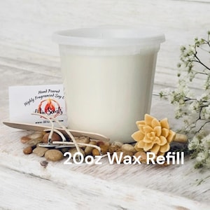 May include: A white plastic container filled with white wax, labeled "Hand Poured Highly Fragranced Soy Candle" with the text "Mix Scents" and a website address. The container is sitting on a wooden surface with a small wooden spoon, a few small stones, and a small, light brown, flower-shaped object. The text "20oz Wax Refill" is written below the container.