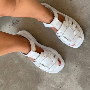 May include: White leather sandals with a chunky platform sole and multiple straps. The sandals have a casual, sporty style.