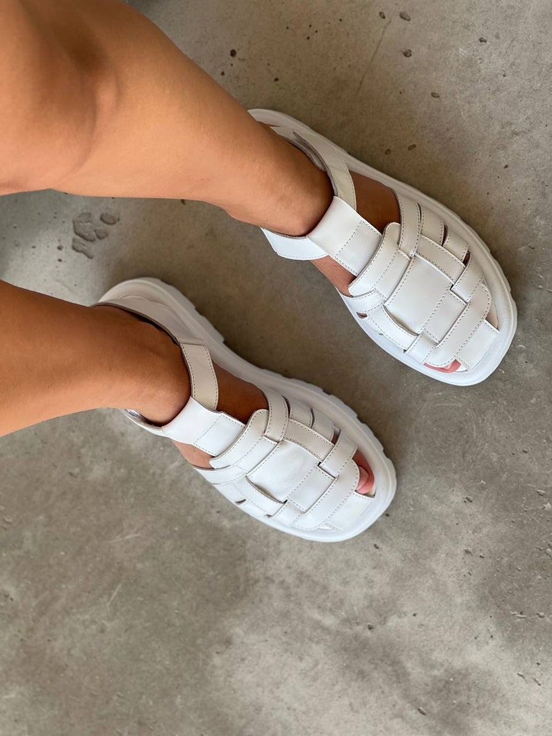 May include: White leather sandals with a chunky platform sole and multiple straps. The sandals have a casual, sporty style.