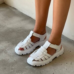May include: White leather sandals with a chunky platform sole and a caged design. The sandals have a velcro strap closure.
