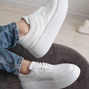 May include: White platform sneakers with a lace-up closure and a thick white sole. The sneakers are made of leather and have a perforated design on the toe.