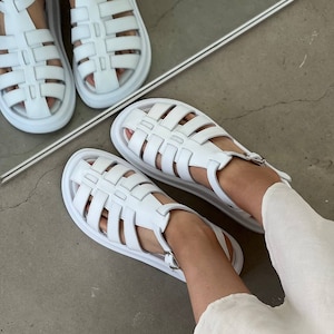 May include: White leather sandals with a cage design and a thick platform sole. The sandals have a buckle closure at the ankle.