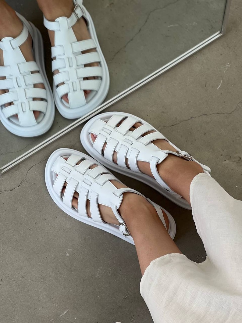 May include: White leather sandals with a cage design and a thick platform sole. The sandals have a buckle closure at the ankle.