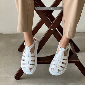 May include: White leather sandals with a thick platform sole and a criss-cross strap design. The sandals have a buckle closure at the ankle.