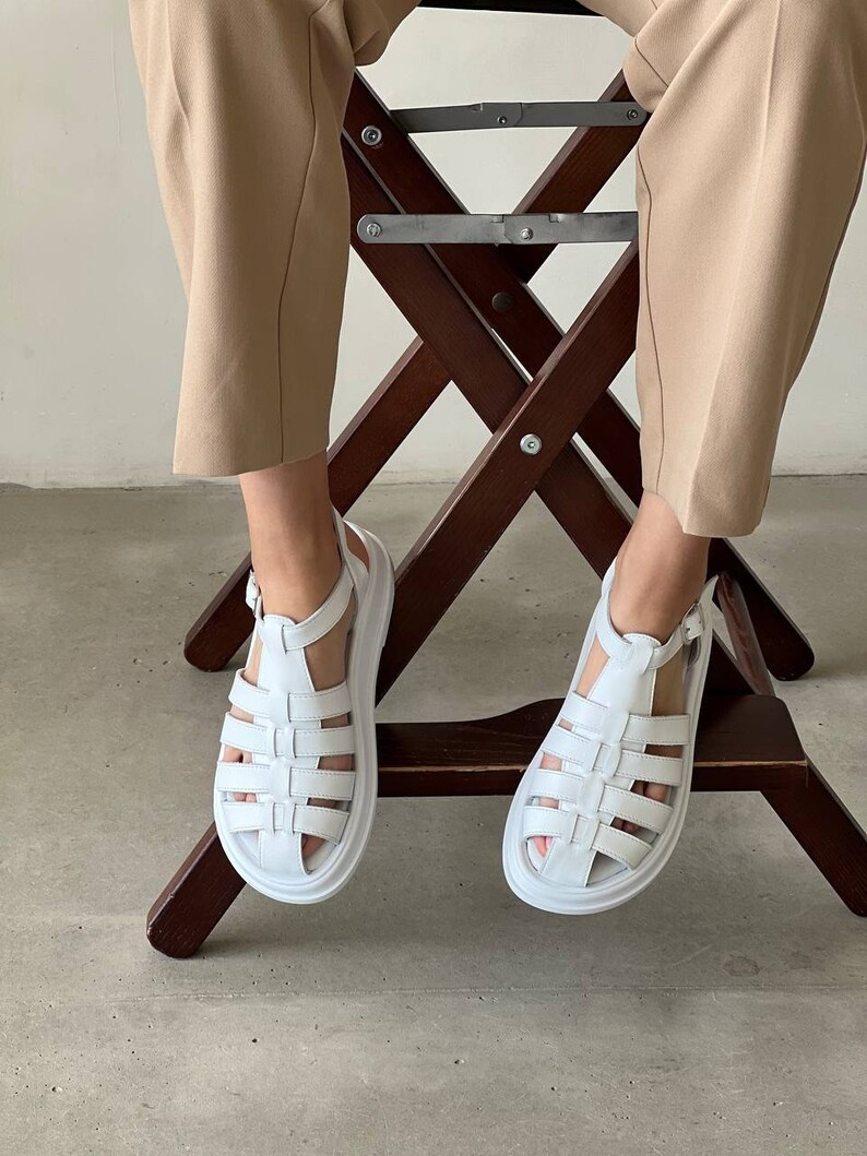 May include: White leather sandals with a thick platform sole and a criss-cross strap design. The sandals have a buckle closure at the ankle.