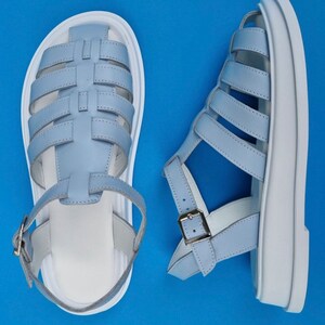 May include: A pair of light blue leather sandals with white soles. The sandals have a criss-cross strap design and a buckle closure.