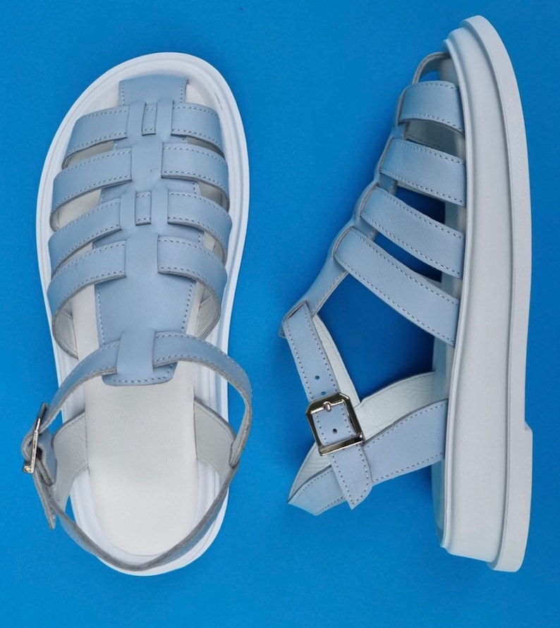 May include: A pair of light blue leather sandals with white soles. The sandals have a criss-cross strap design and a buckle closure.