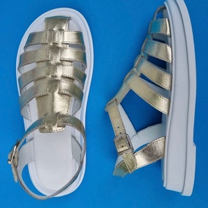 May include: A pair of gold leather sandals with white soles and straps. The sandals have a caged design and a buckle closure.