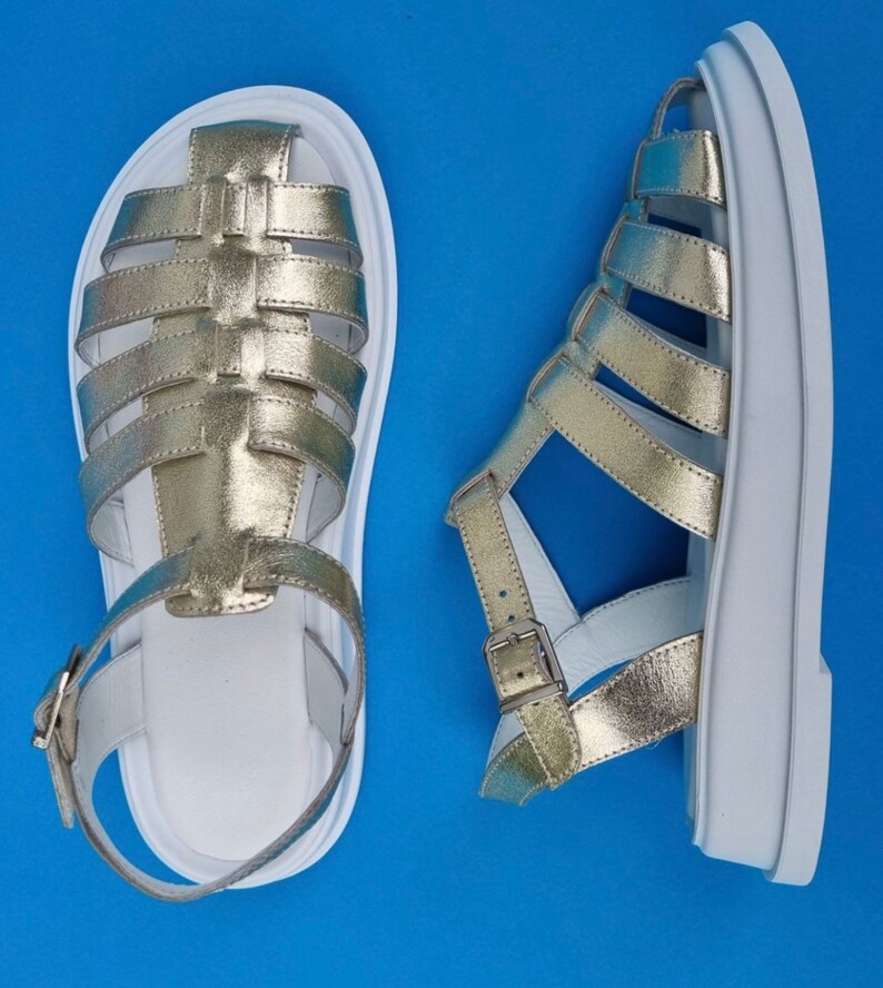 May include: A pair of gold leather sandals with white soles and straps. The sandals have a caged design and a buckle closure.