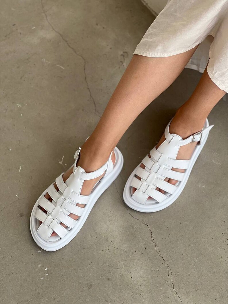 May include: White leather sandals with a chunky platform sole and a cage-like design. The sandals have a buckle closure at the ankle.