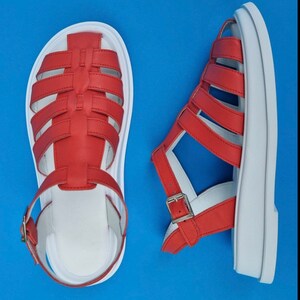 May include: A pair of red leather sandals with white soles. The sandals have a criss-cross strap design and a buckle closure.