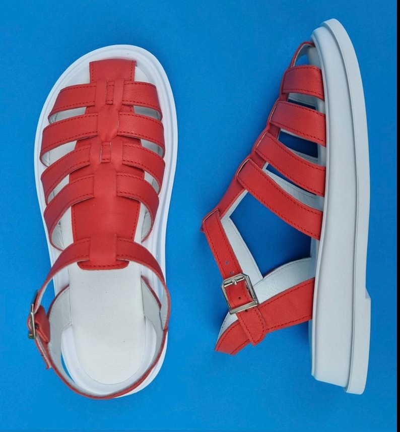 May include: A pair of red leather sandals with white soles. The sandals have a criss-cross strap design and a buckle closure.