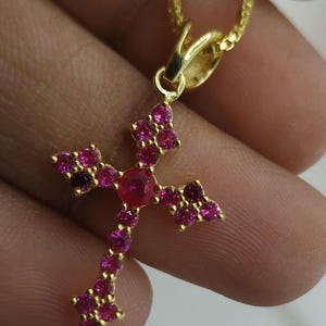 May include: A gold-colored cross pendant necklace with a chain. The cross is adorned with numerous pink and red gemstones. The pendant has a central, larger red gemstone, and smaller pink gemstones form the cross's arms.