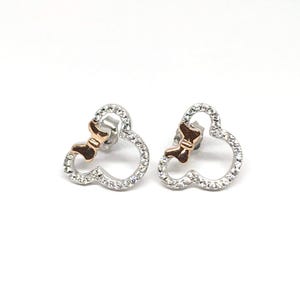 May include: A pair of silver stud earrings. Each earring is shaped like a mouse head with a bow. The mouse head outline is encrusted with small, clear crystals. The bow is a rose gold colour. The earrings are on a white background.